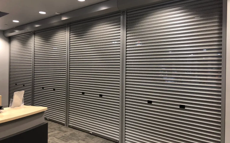 Exterior of a retail store with closed metal security shutters covering windows and entrance, framed by brick and stucco facade.